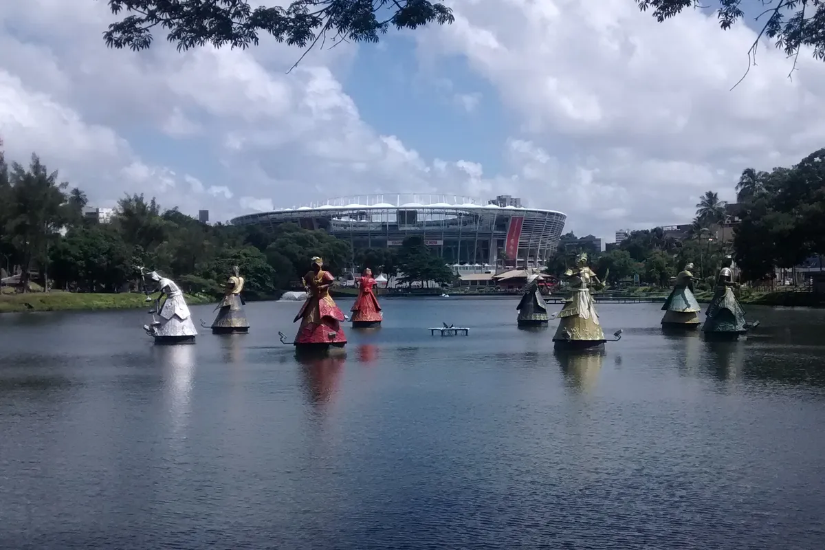 Arena Fonte Nova stadium in Salvador seen across the Dique do Tororó lagoon, with colorful Orixá sculptures standing in the water