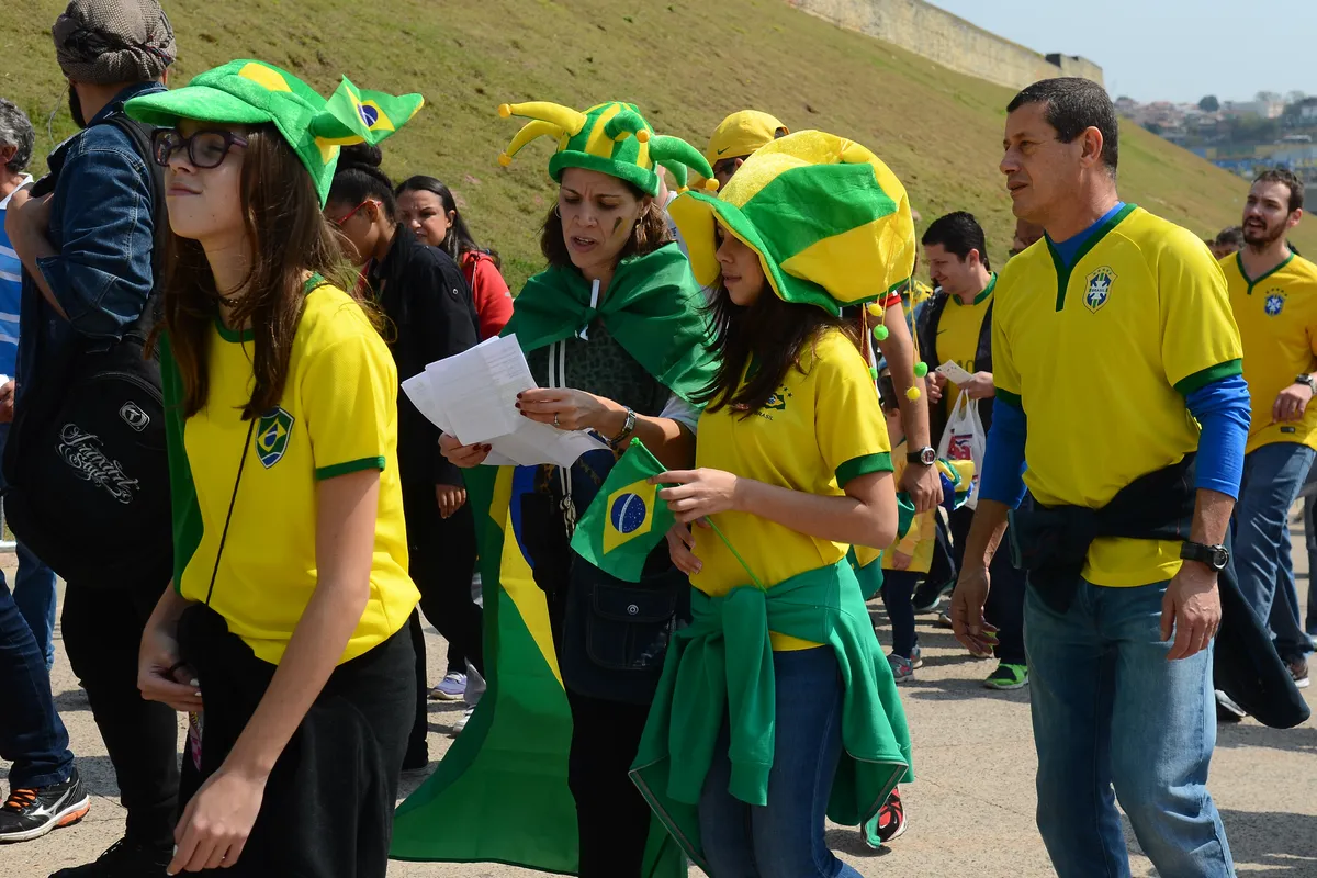 Brazil fans in yellow shirts, jester hats and a flag walk toward Arena Corinthians in São Paulo, one woman reading a printed ticket