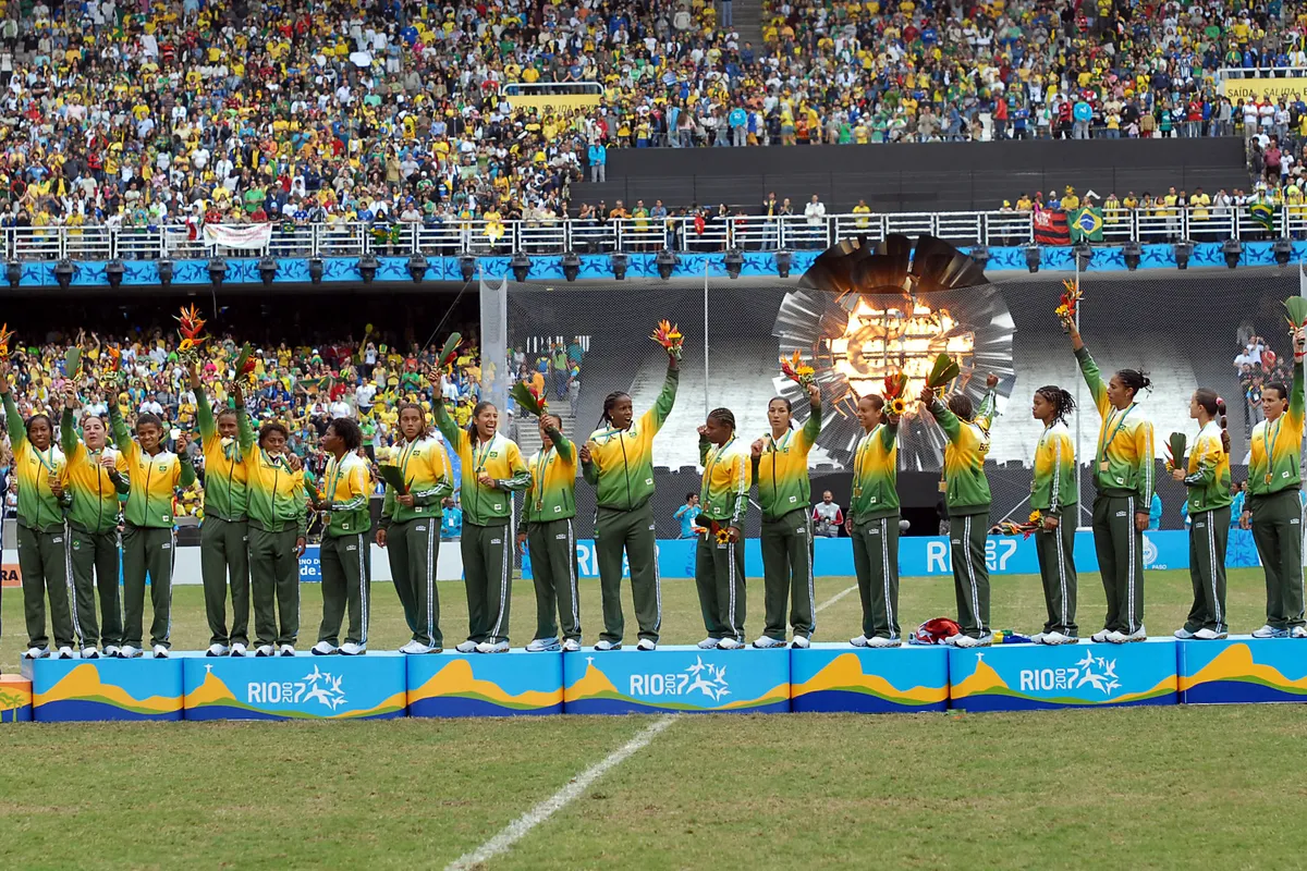 Brazil women's soccer players on a podium in green and yellow tracksuits with arms raised, holding flowers and gold medals, in front of a packed stadium