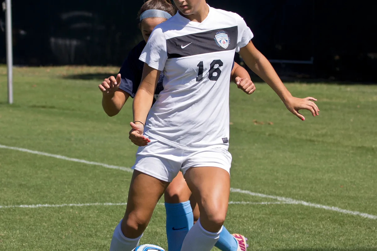 A college women's soccer player in a white jersey number 16 and a light blue headband shields the ball from a defender on a grass field