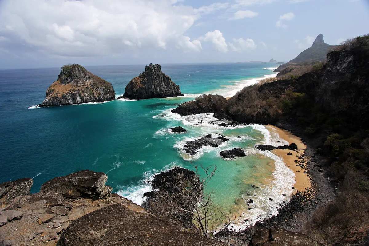 Turquoise water in Baía dos Porcos on Fernando de Noronha, with the two rocky peaks of Morro Dois Irmãos rising from the sea and a curved beach below a green headland