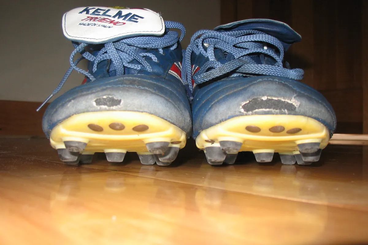 Front view of a pair of blue soccer cleats on a wooden floor, showing the molded studs under the toe of each shoe