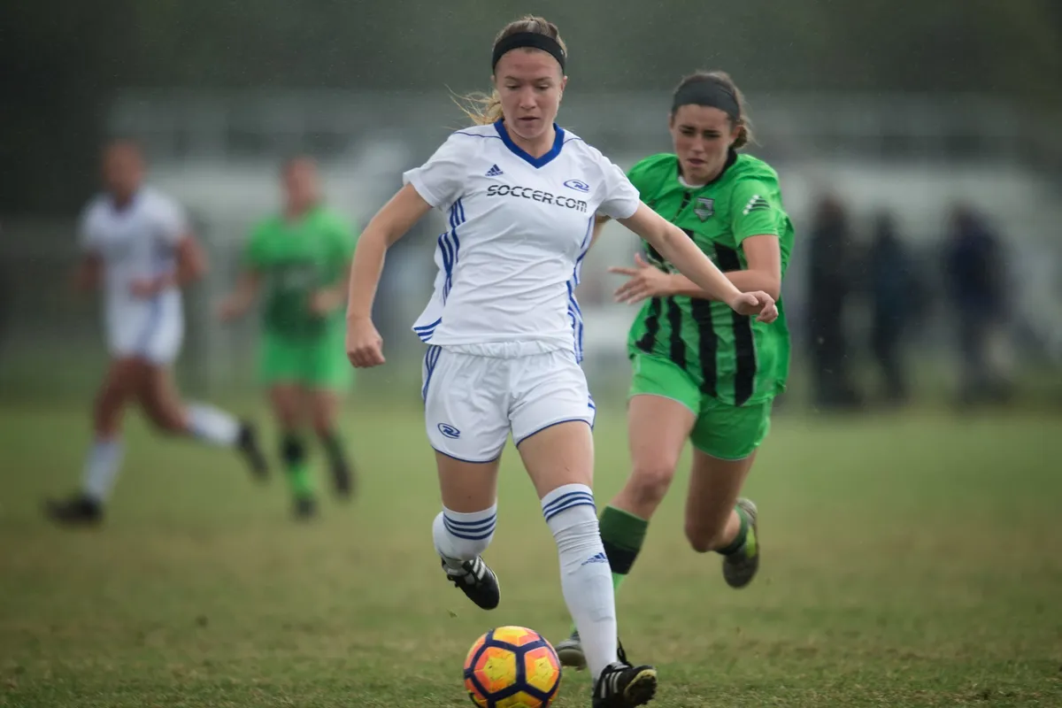 A girl in a white club soccer jersey dribbles the ball in light rain while a defender in a green jersey chases her, with other players blurred behind