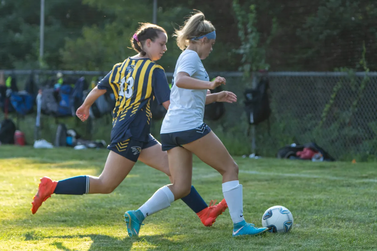Two girls race for the ball in a soccer game, one in a navy and gold striped jersey and one in a white shirt with a blue headband