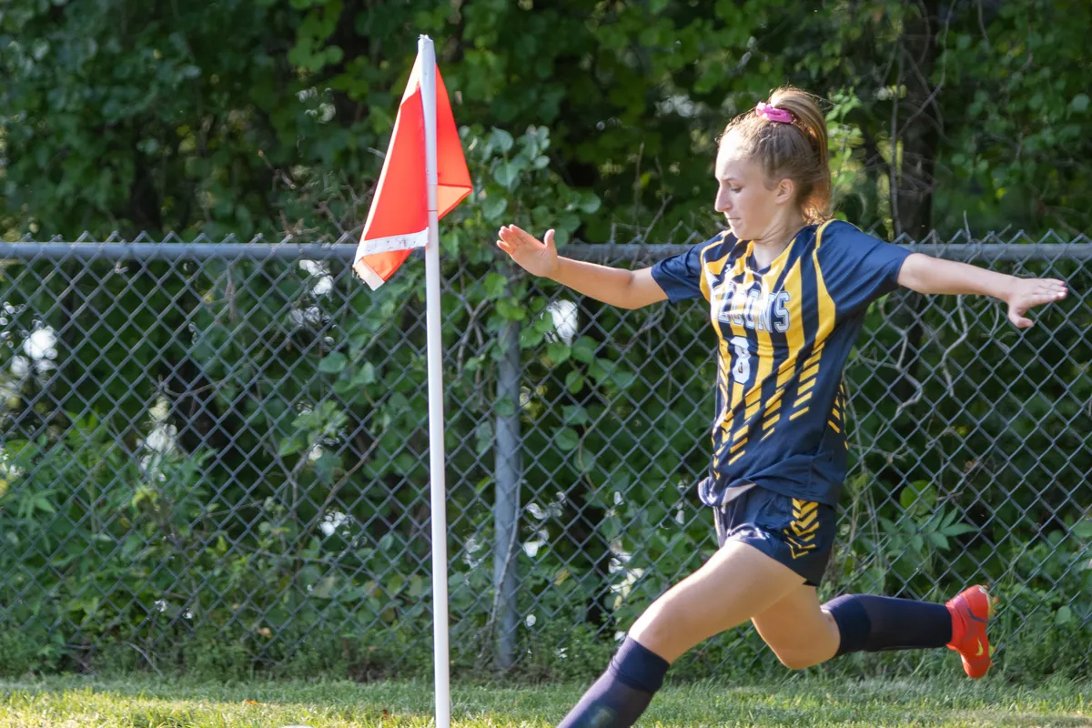 A girl in a navy and gold striped jersey swings her leg to take a corner kick beside the orange corner flag