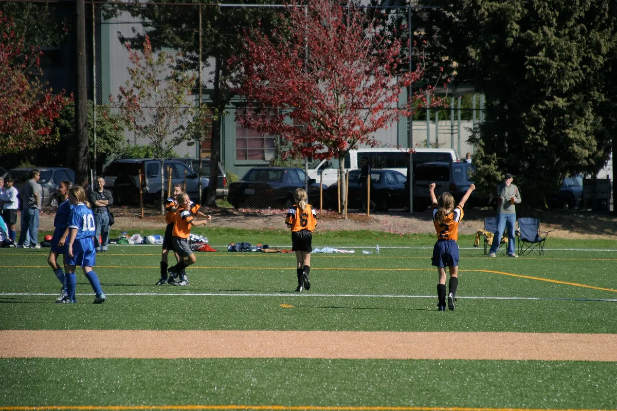 Girls in orange jerseys celebrating on a youth soccer field, one with both arms raised, while parents watch from folding chairs near the sideline