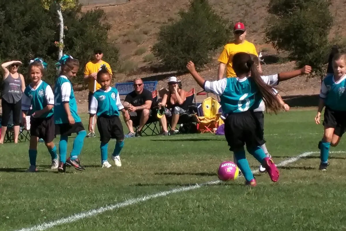 Young girls in teal and white uniforms playing a youth soccer game on a grass field, one girl about to kick a pink ball while parents watch from folding chairs