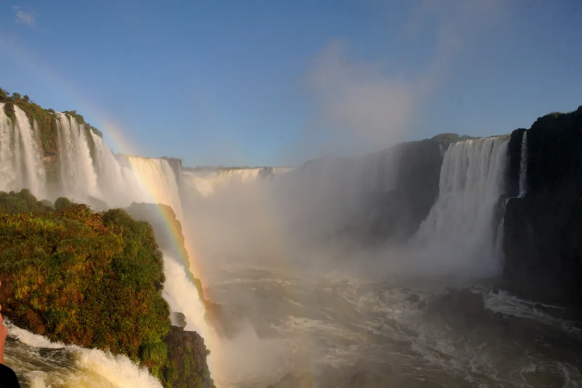 Water thundering over the Devil's Throat at Iguazu Falls, seen from the Brazilian side, with a rainbow in the mist and forest on the cliffs