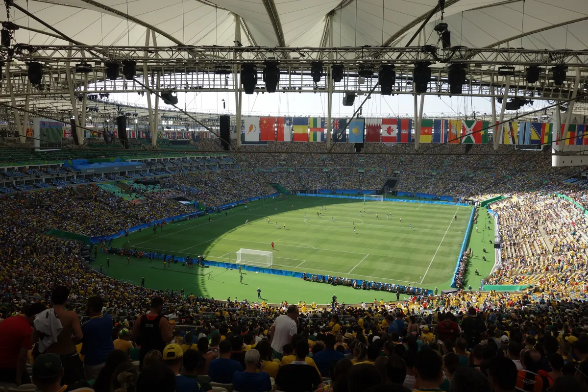 Inside the Maracanã stadium during a football match, with packed yellow and green stands, a roof of white fabric and steel beams, and a row of national flags above the pitch