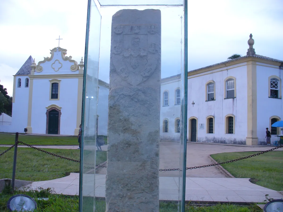 The Marco do Descobrimento, a weathered stone marker carved with the arms of Portugal, inside a glass case in Porto Seguro's Historic City, with a white and yellow colonial church and a long white building behind it