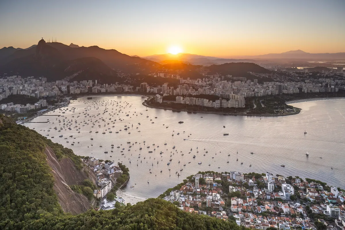 Sunset over Botafogo Bay in Rio de Janeiro, with sailboats on the water, apartment buildings along the shore and the Christ the Redeemer statue on a mountain in the background