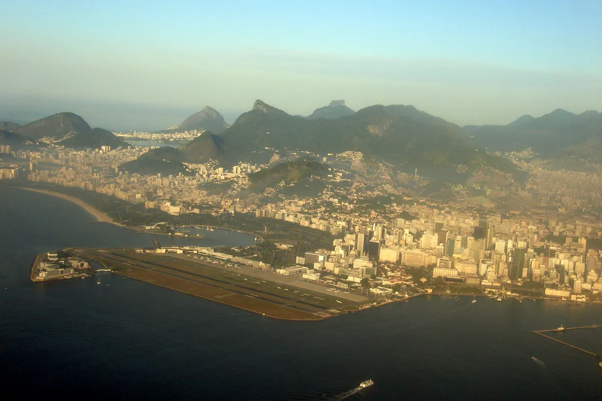 Aerial view of the Santos Dumont airport in Rio de Janeiro, a long runway on a strip of land beside the bay, with downtown buildings and green mountains behind it