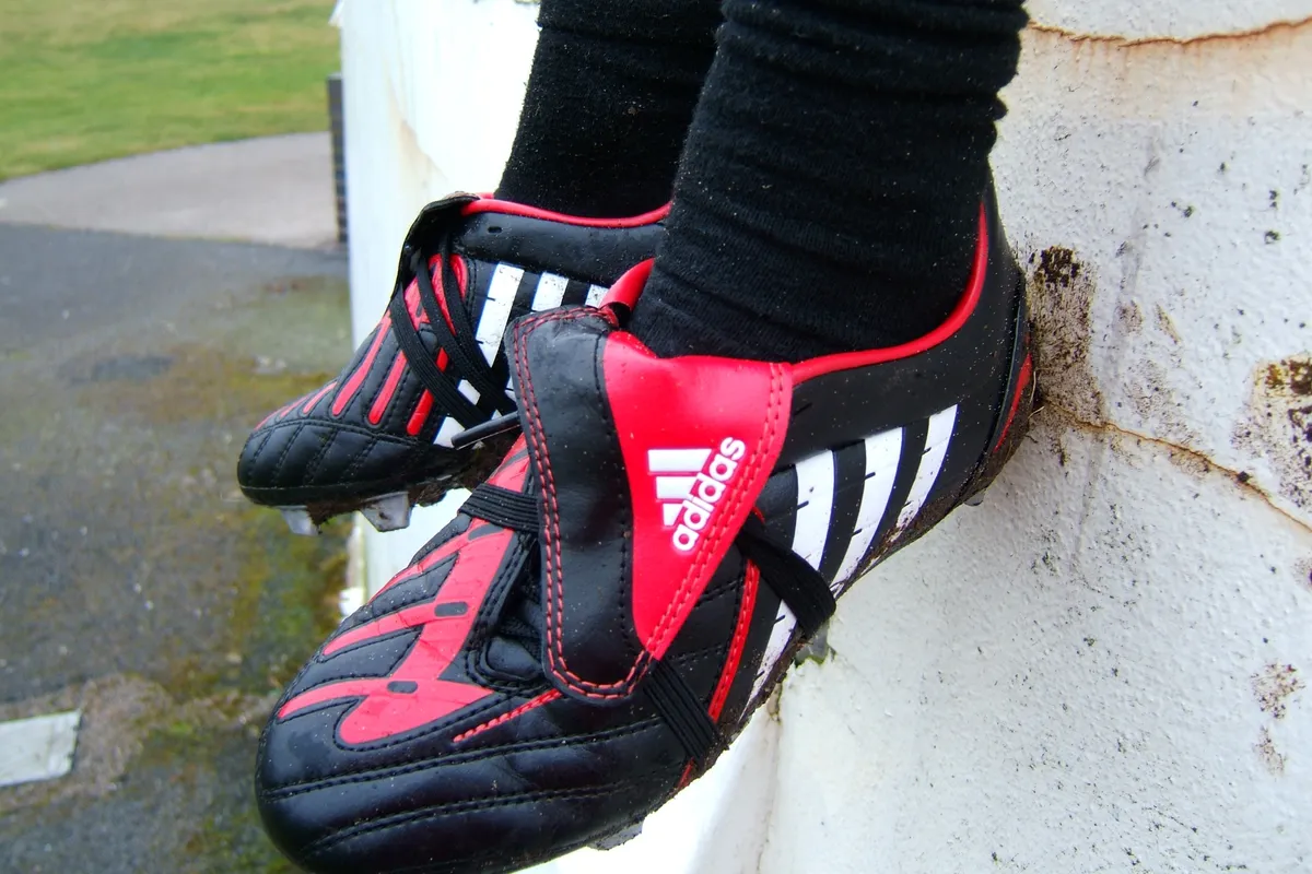 A person's feet in black socks and black, red and white soccer boots, resting against a white wall with grass in the background