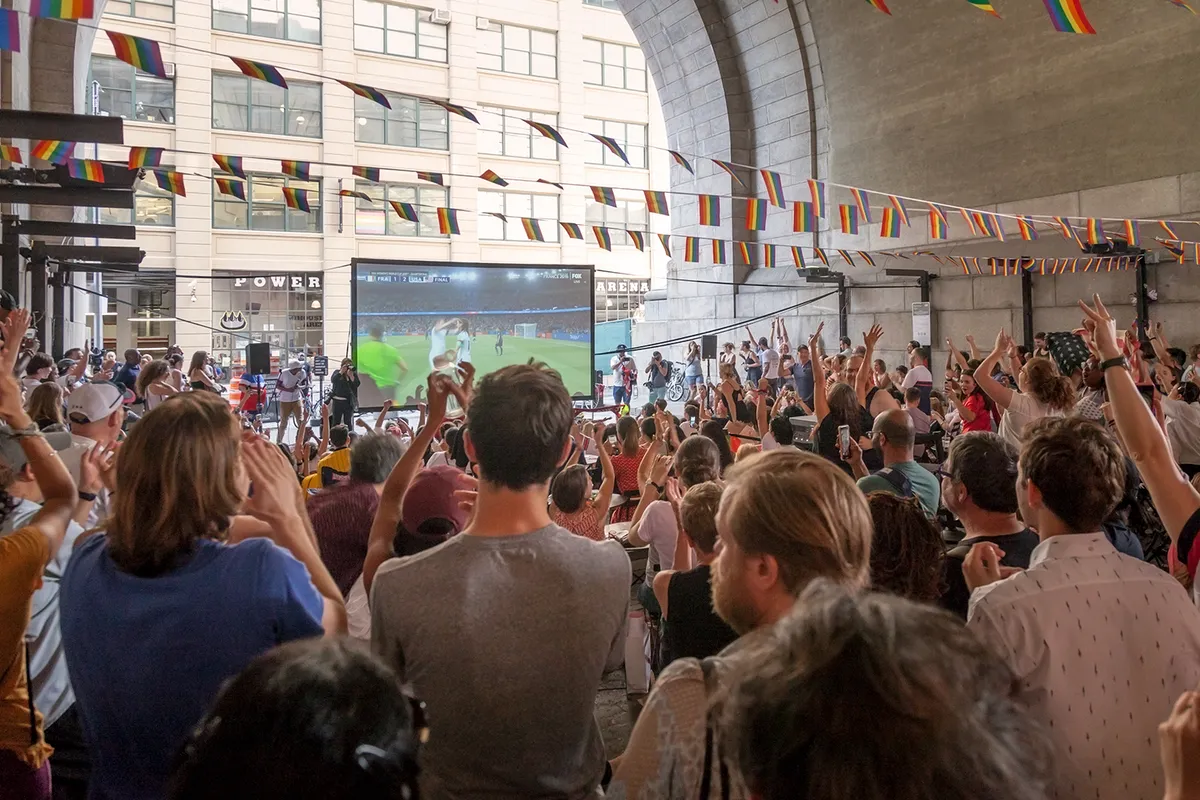 Fans raising their arms and cheering in front of a big outdoor screen showing a Women's World Cup goal celebration
