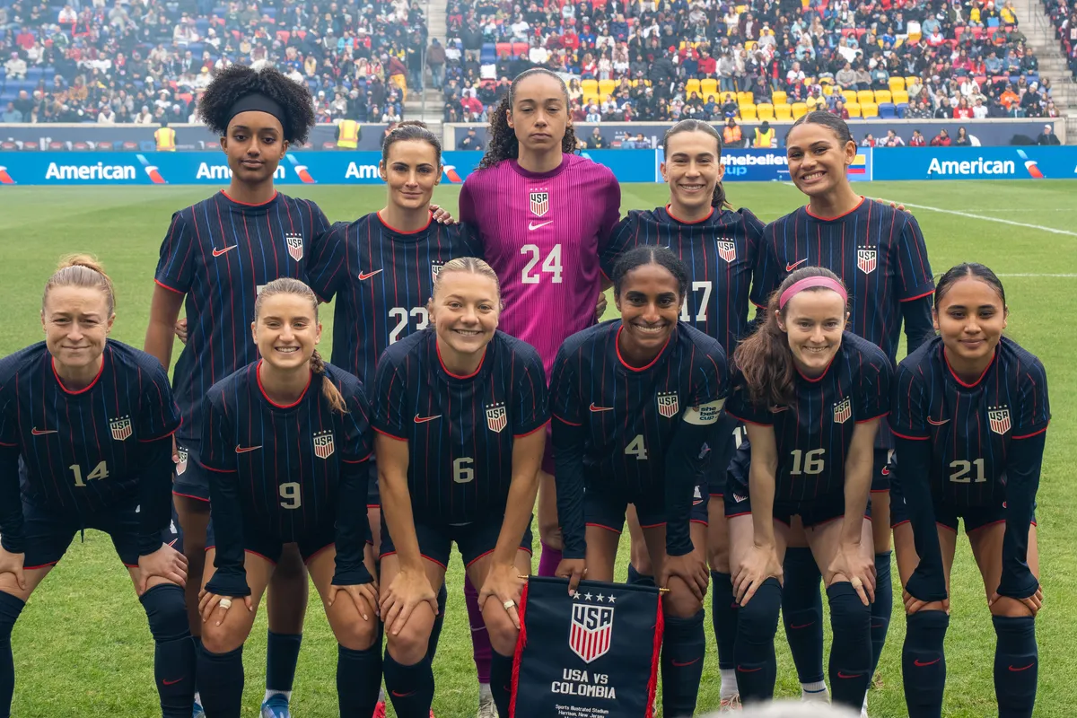 The U.S. women's national team starting eleven in navy jerseys pose in two rows on the field before a match, with a USA v Colombia pennant