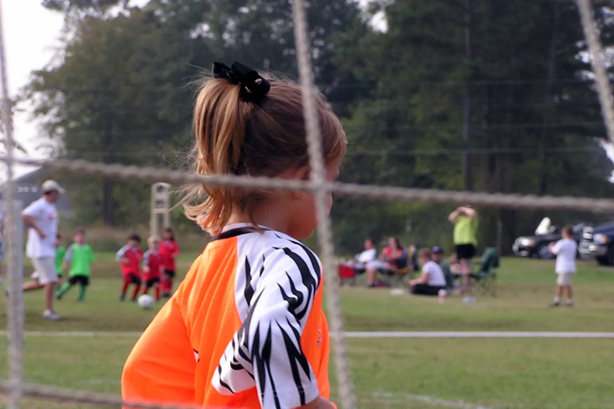 A young girl in an orange jersey stands behind a soccer net watching a recreational game, with other players and parents in folding chairs on the field behind her